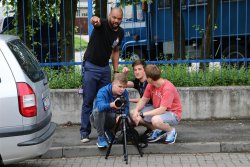 Troje uczestników warsztatów wraz z opiekunem realizujący zdjęcia do filmu na chodniku, w tle płot z policyjnym samochodem ciężarowym.